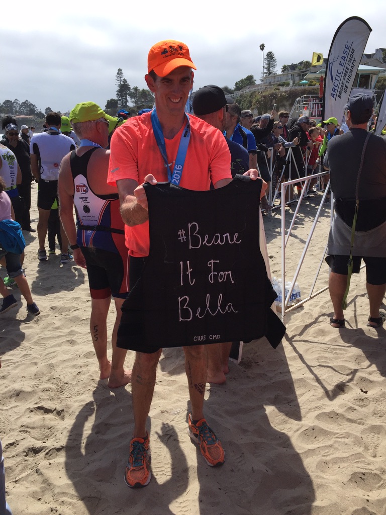 Mark at the 2016 Half Ironman finish line, holding his weighted vest reading #Beare It For Bella · Cure CMD